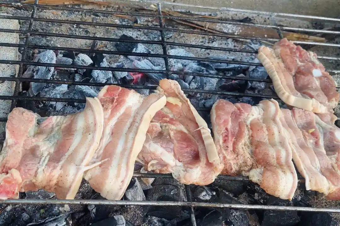 Strips of pork belly grilling over charcoal coals on a beachside grill in Jomalig