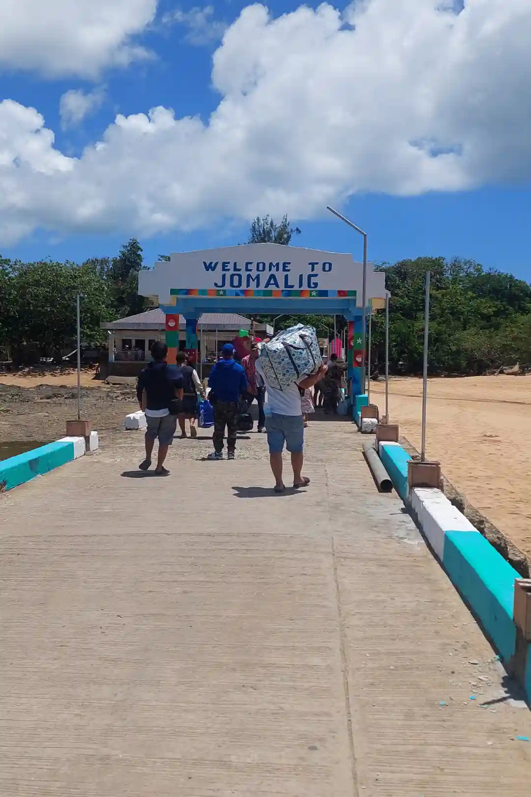 Travelers carrying luggage past the Welcome to Jomalig sign at the island port entrance