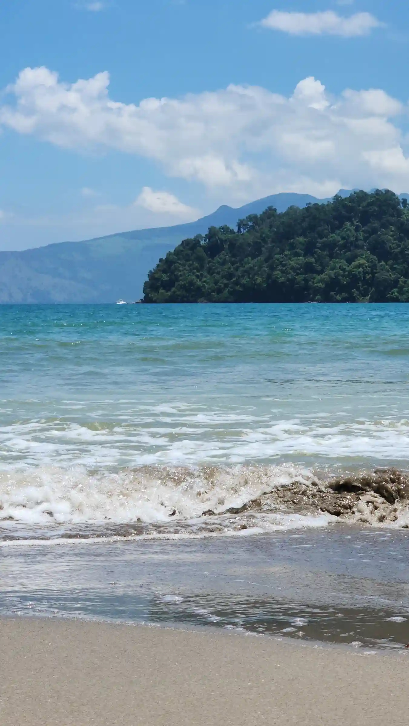 Turquoise waves rolling onto a quiet sandy shore with a forested headland and distant mountains in Morong Bataan