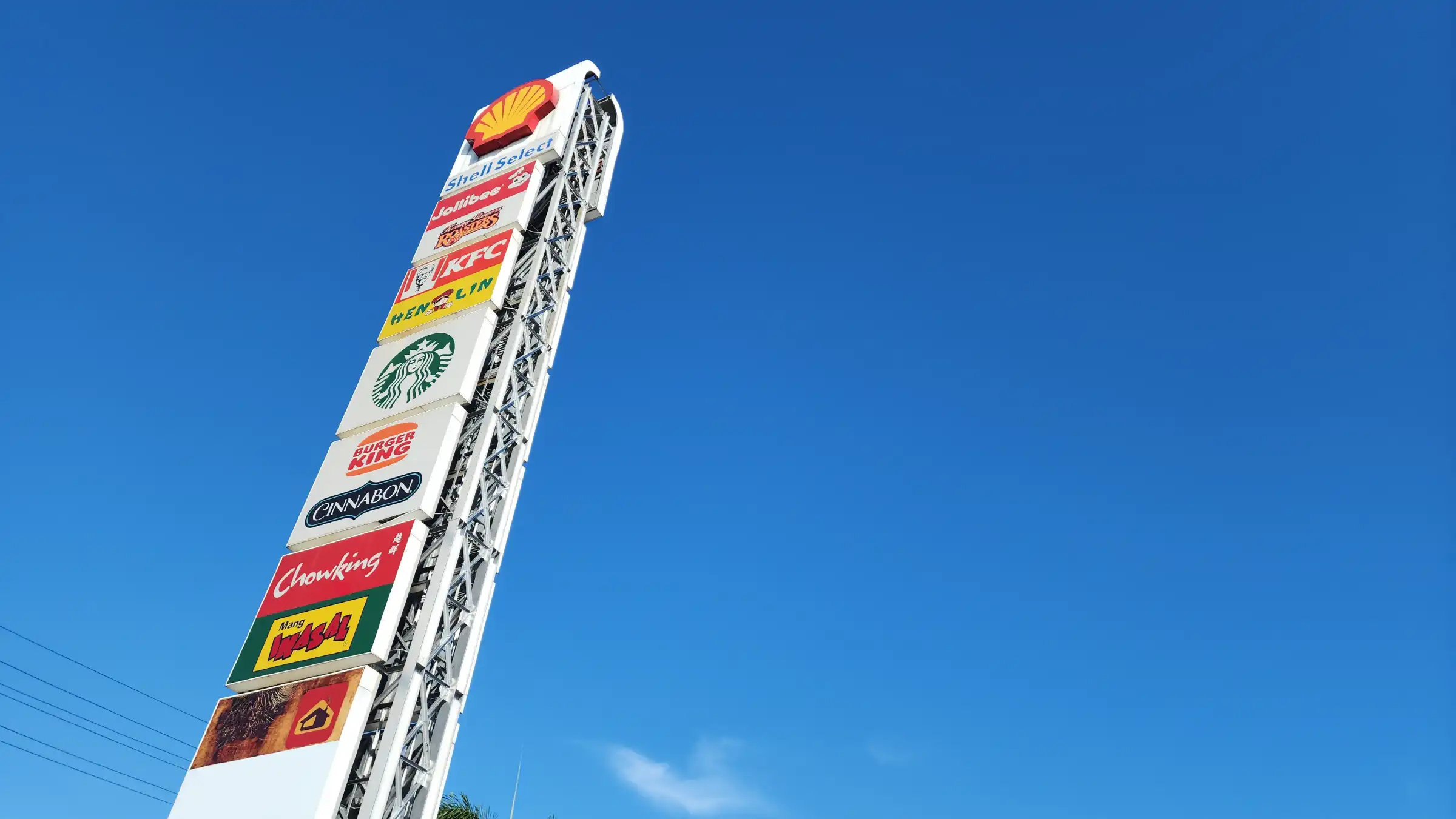 Tall roadside signage tower of food brands against a clear blue sky at a Shell station in Balagtas
