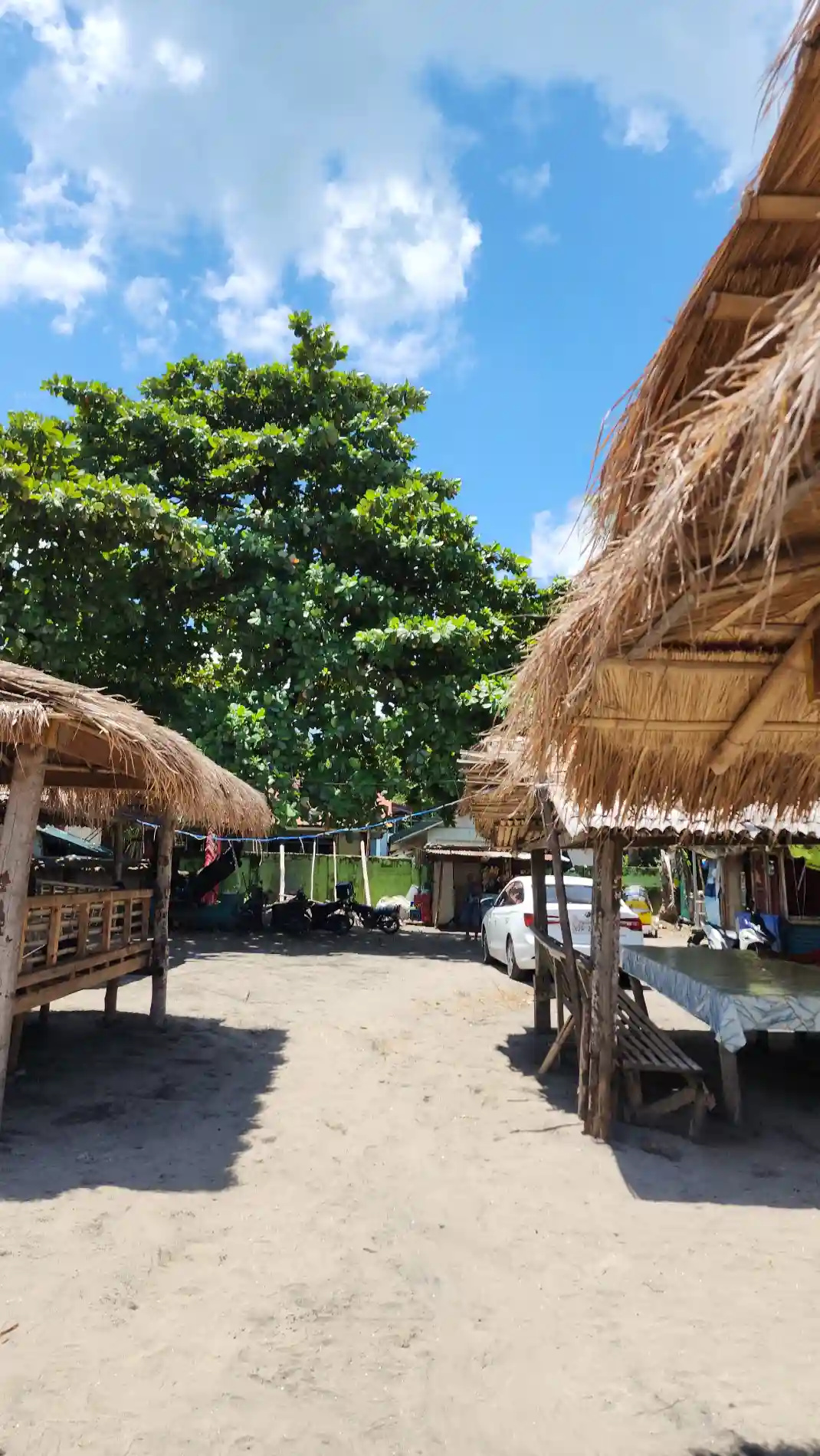 Nipa roofed cottages and parked motorcycles under a large tree in a sandy beachfront clearing in Morong, Bataan