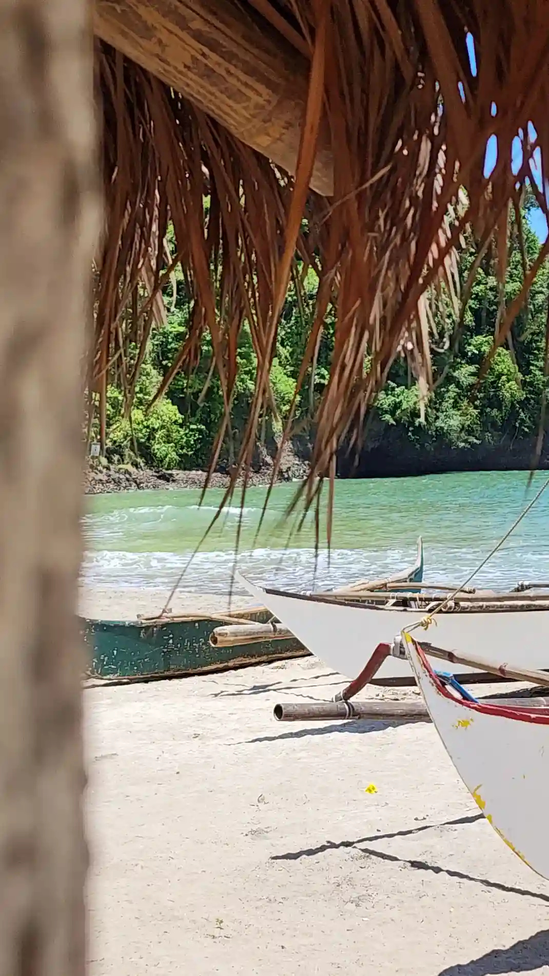 Outrigger boats on the sand seen through a cogon roof edge with green water and forested hills in Morong, Bataan