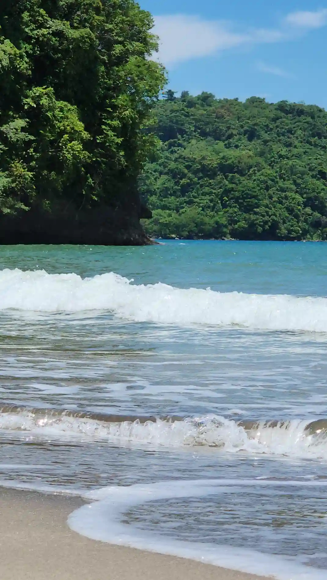 Turquoise waves washing over open sandy shore with a forested point and mountain backdrop at a beach in Morong, Bataan