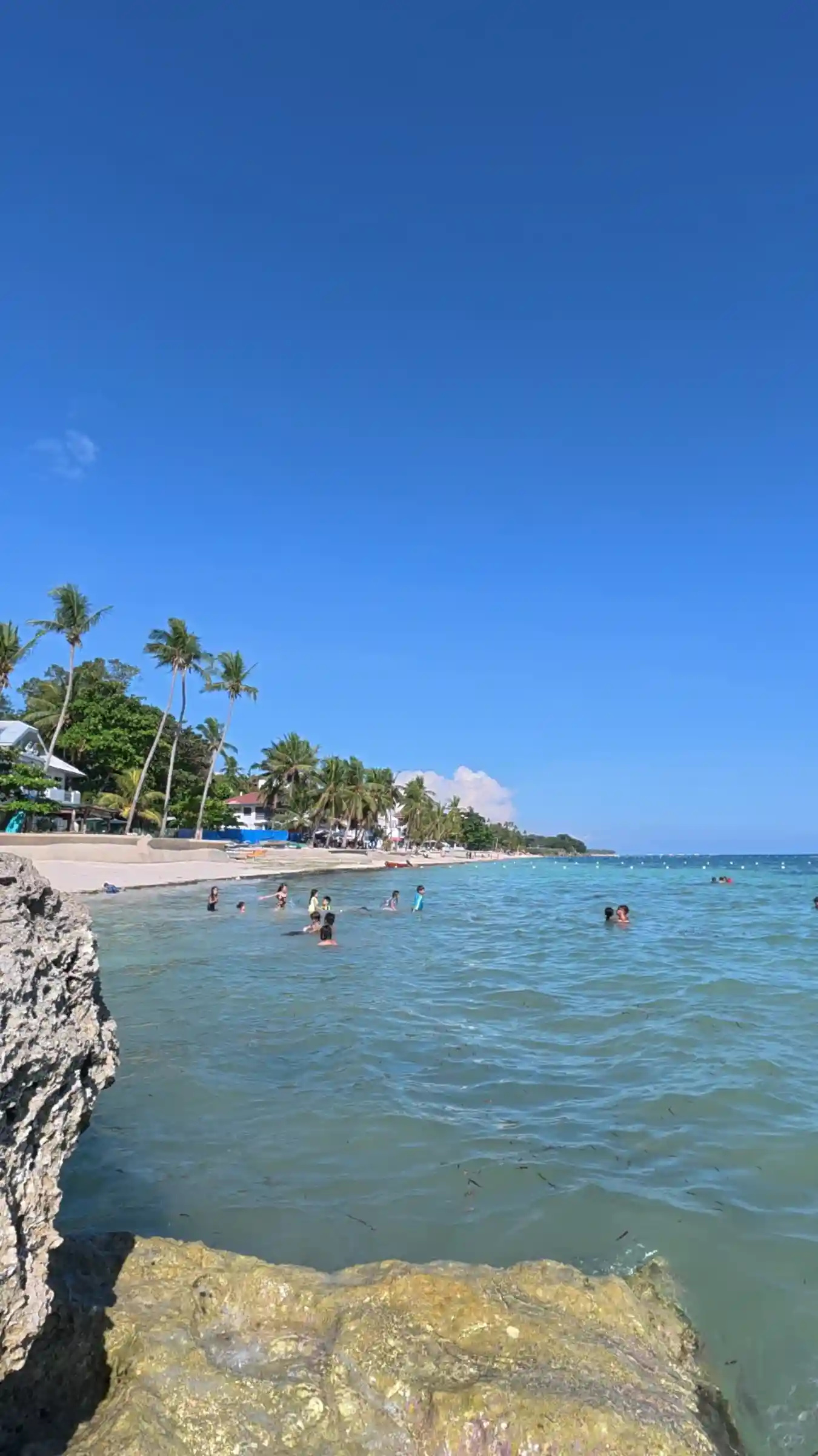 Large rock formation beside turquoise water with swimmers and coastal resorts along Kalipayan Beach Panglao