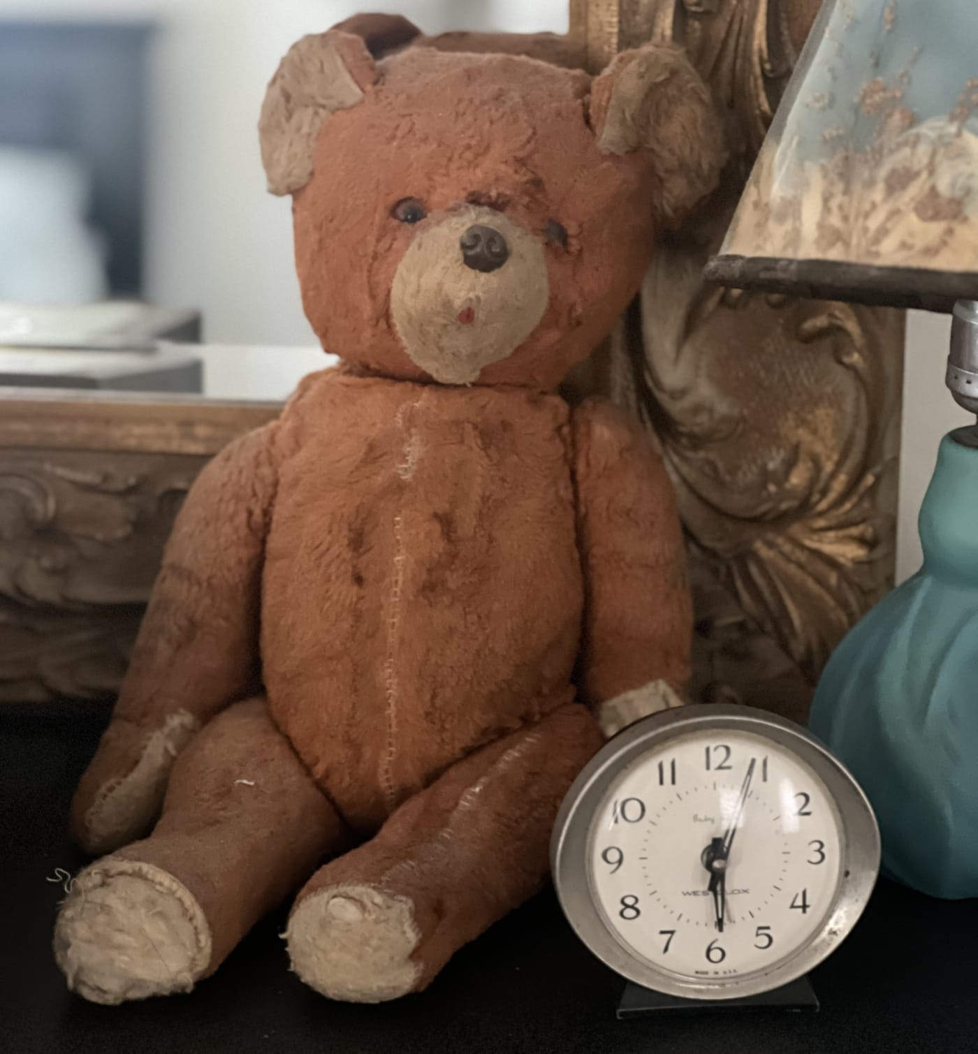 A well-worn brown teddy bear sitting on a dark tabletop beside a small silver Westclox alarm clock, in front of a gilded mirror frame.