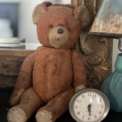 A well-worn brown teddy bear sitting on a dark tabletop beside a small silver Westclox alarm clock, in front of a gilded mirror frame.