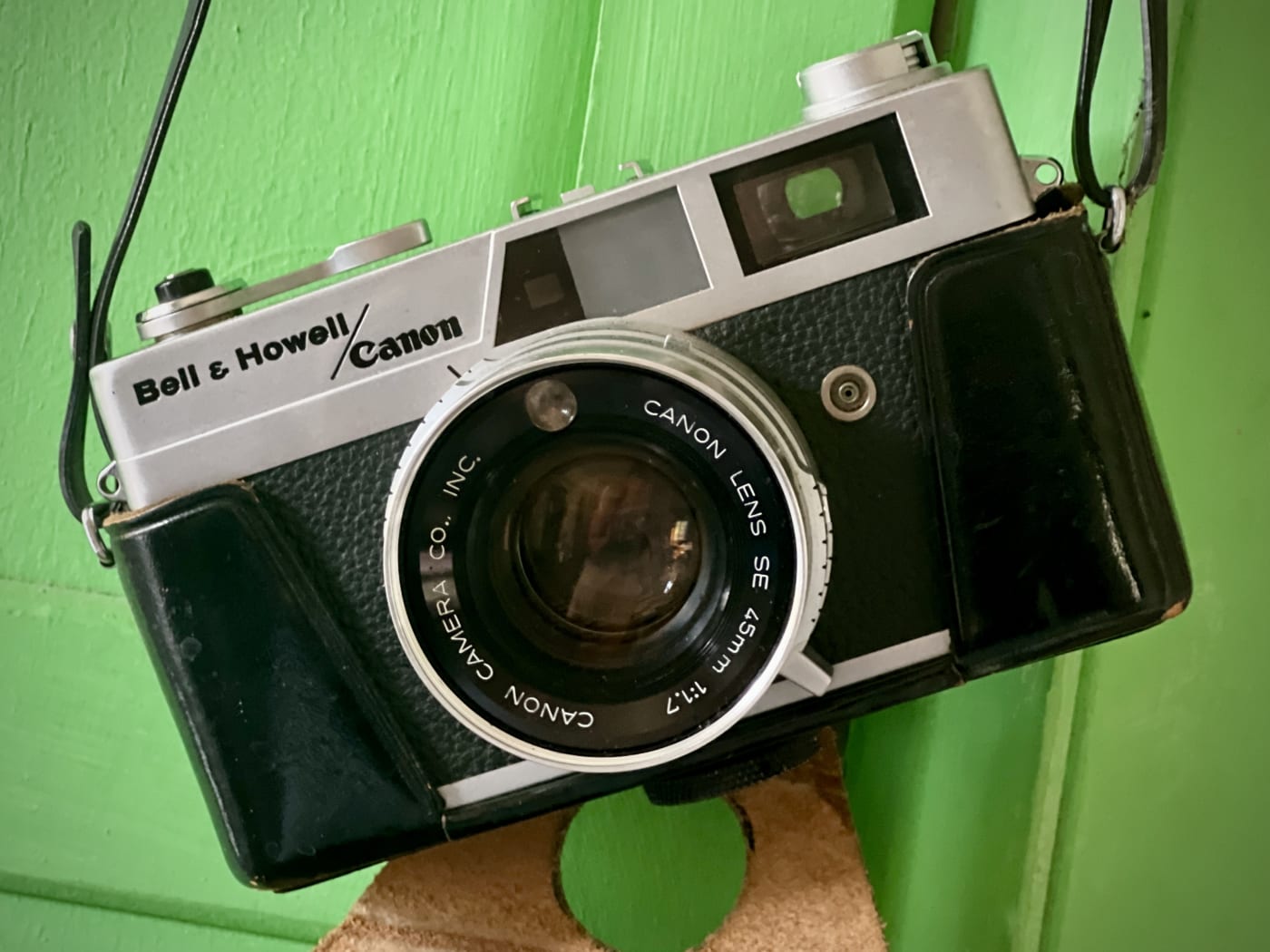 A vintage Bell & Howell/Canon 35mm rangefinder camera in a worn black leather case, hanging by its strap against a bright green wooden wall.