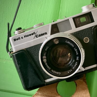A vintage Bell & Howell/Canon 35mm rangefinder camera in a worn black leather case, hanging by its strap against a bright green wooden wall.