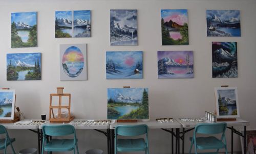 Many landscape paintings hanging on the wall, with painting stations set up at on a table