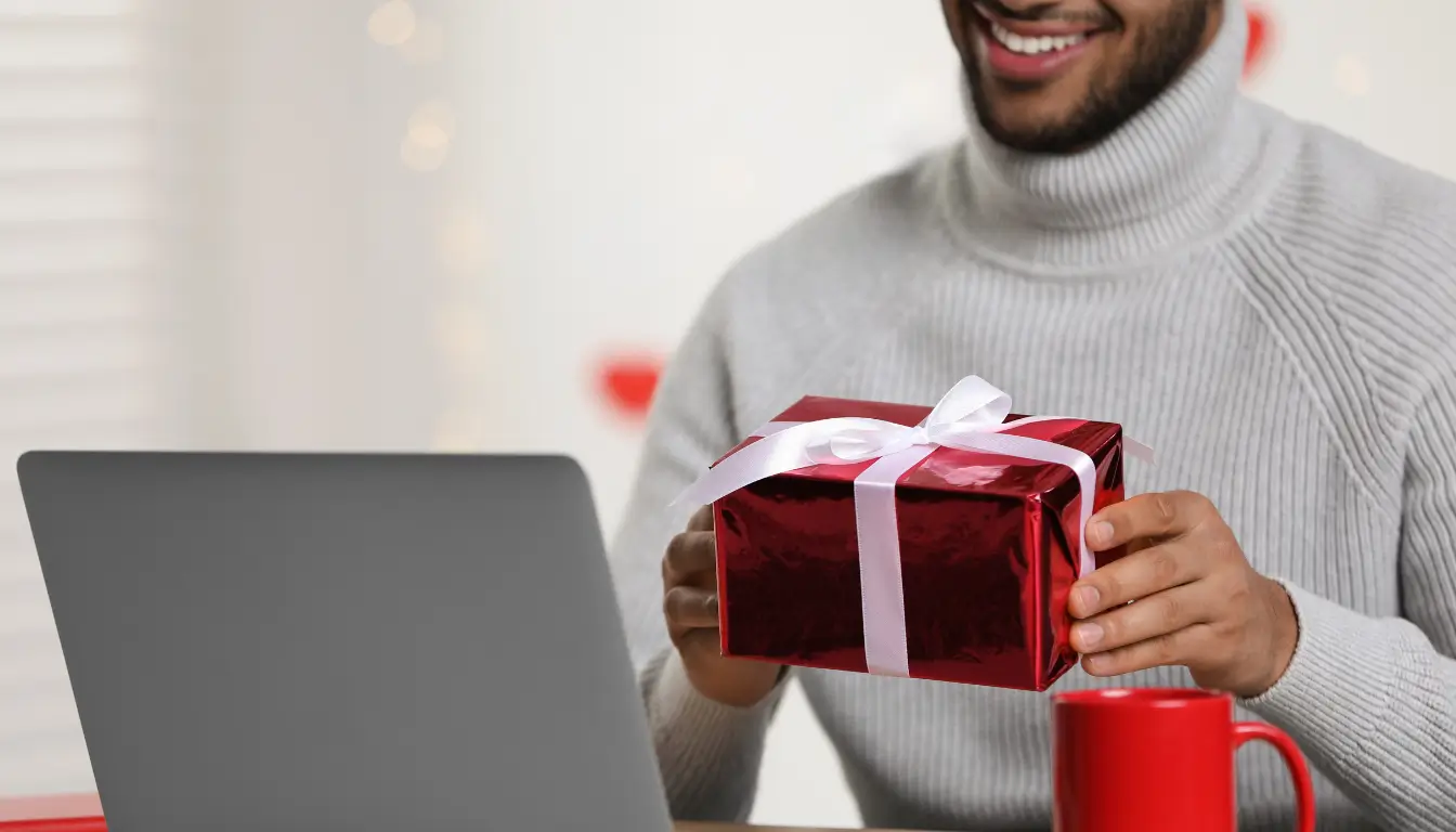 A man smiling while opening a gift on a video call, symbolizing the joy of receiving a care package in a long-distance relationship.