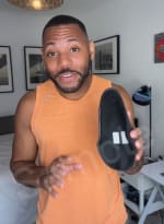 Person in orange tank top holding a black shoe in a room with framed pictures.