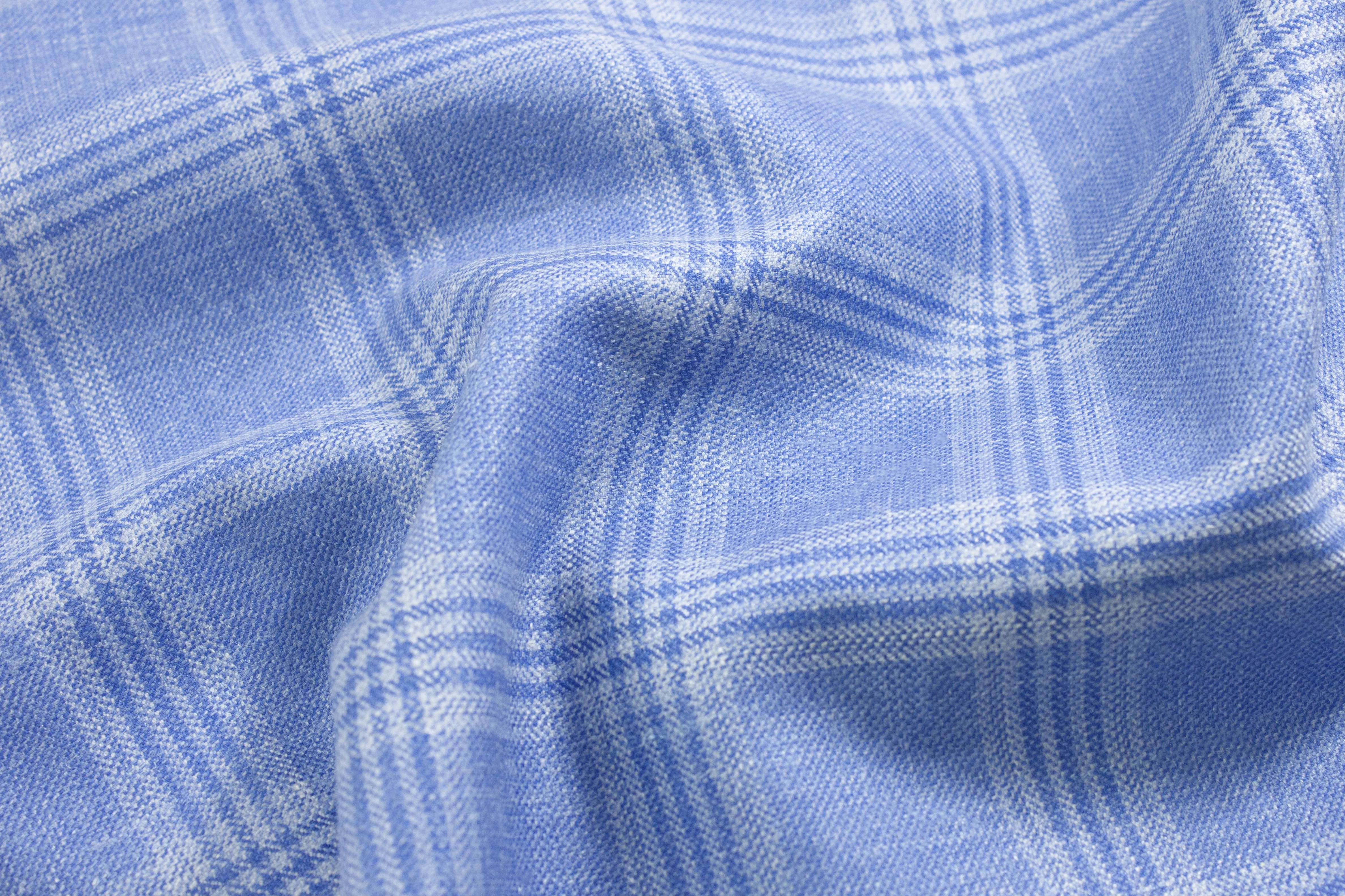 A draped view of the Azure Riviera fabric, highlighting the elegant luster of silk and the breathable weave of linen. The soft blue hues and crisp check pattern suggest a premium, bespoke summer sport coat.