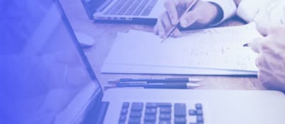 A hand holding a pen on paper next to open laptop