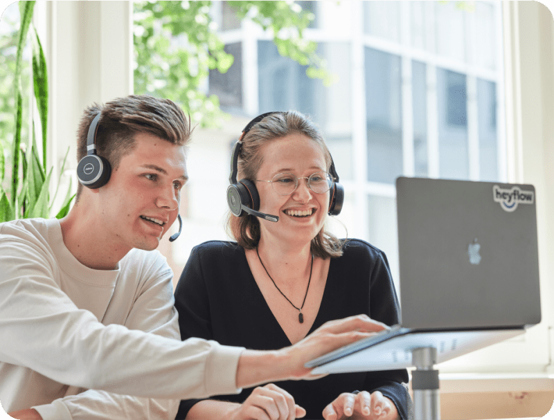 A young male and female caucasian persons with headsets smiling and looking at laptop that has sticker saying Heyflow.