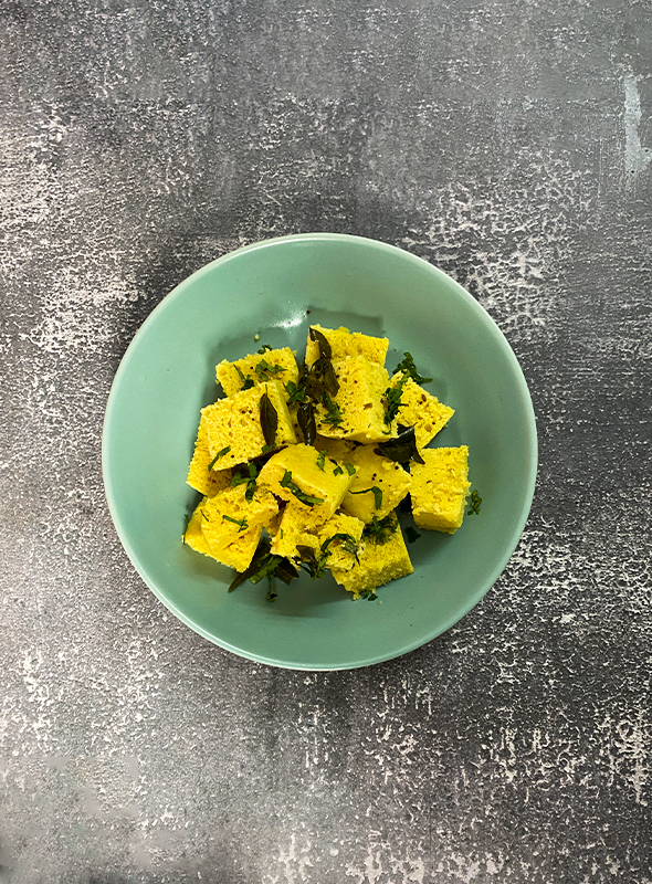 Khaman dhokla is a popular Gujarati snack made from fermented gram flour batter. The batter is spiced with green chilies, ginger, and turmeric, then steamed until light and fluffy. Once cooked, it is tempered with mustard seeds and curry leaves for added flavor