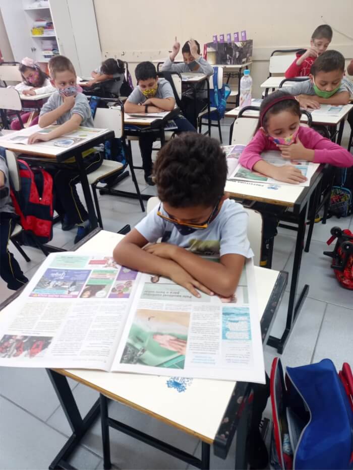 Crianças em suas classes escolares lendo o jornal Nosso Bem Estar