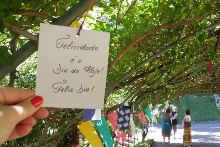 Túnel de plantas com mensagens e mão segurando bilhete Felicidade é o Dia de Hoje - Feliz Dia