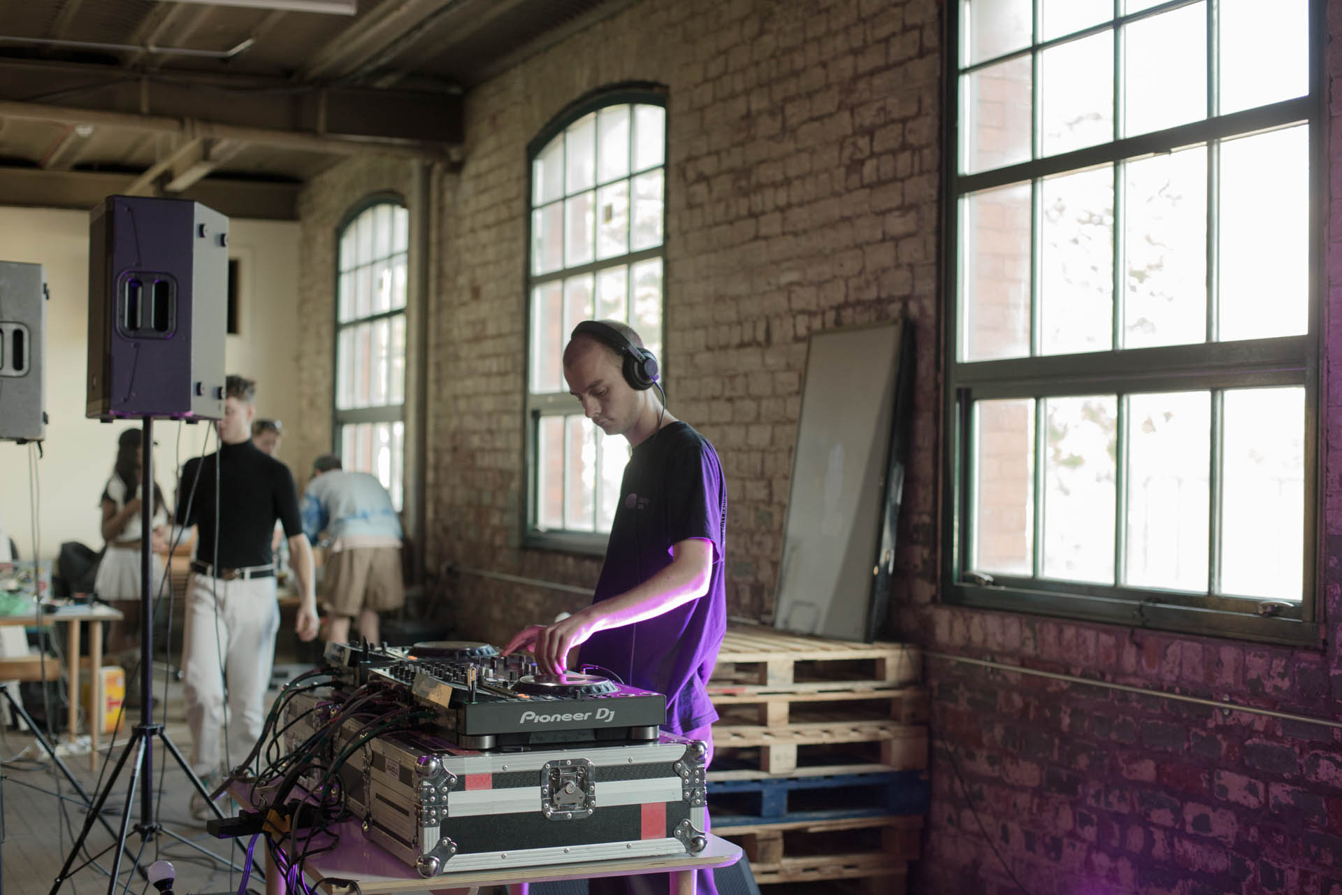 Man DJs in a warehouse looking room, with tech people behind him