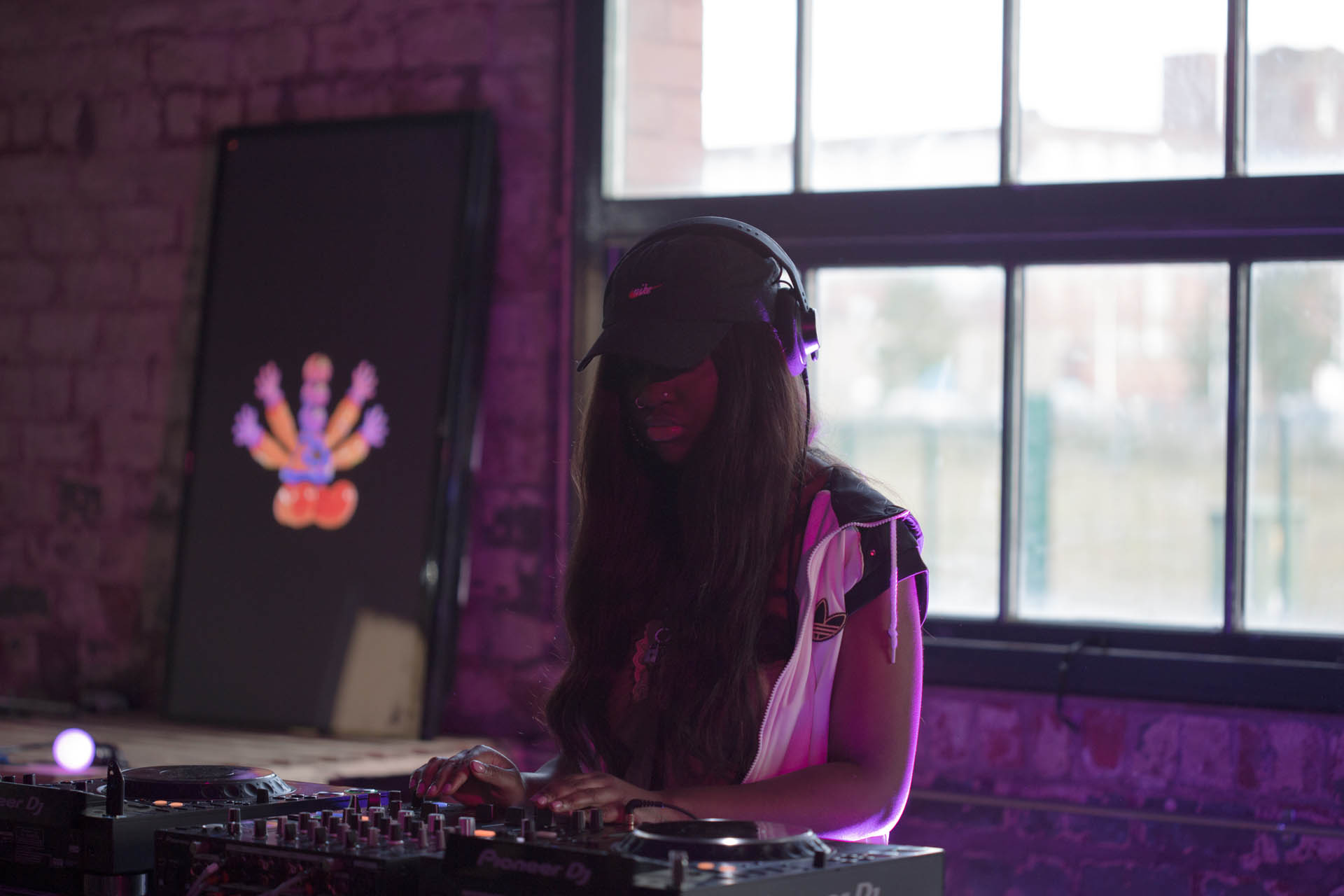 Woman with black cap on DJs, with TV behind her with the GUTS character