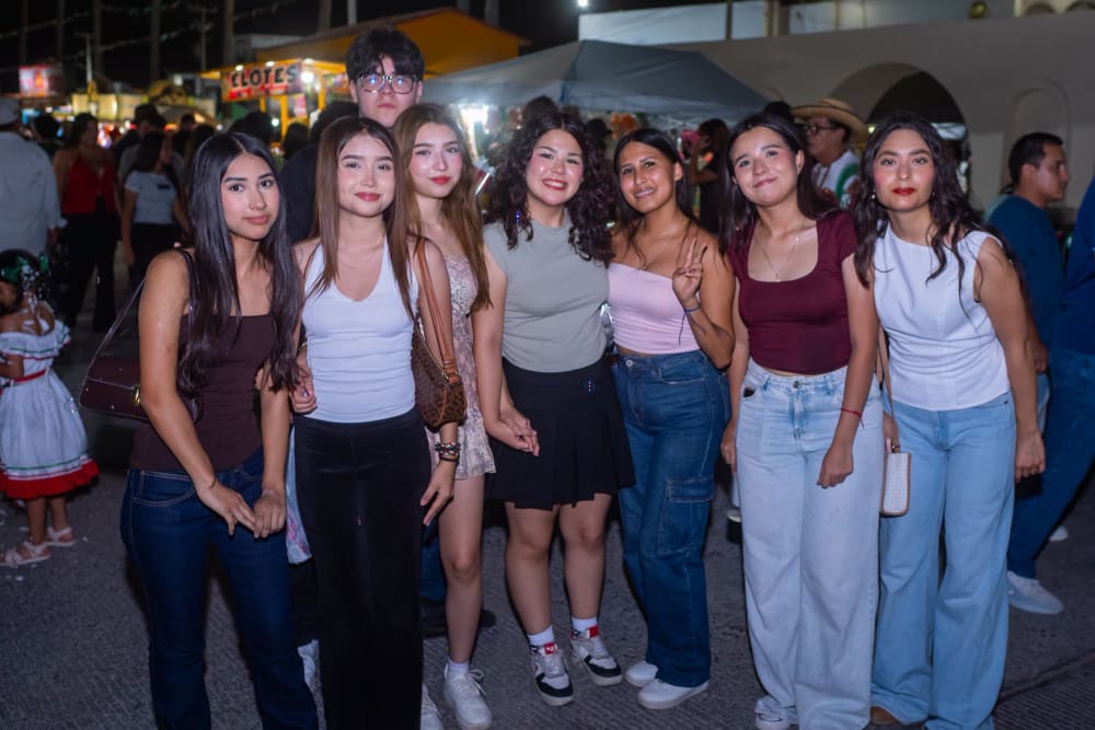 Grupo de jóvenes posando en la kermés frente al puesto de elotes, con una niña de traje folclórico al fondo