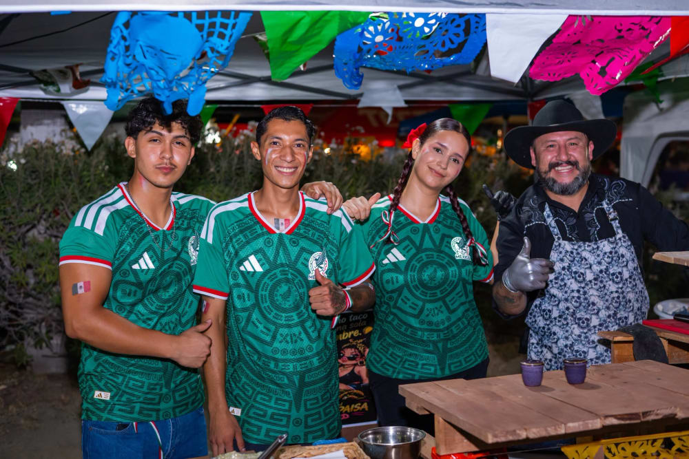 Cuatro personas con la camiseta de la Selección Mexicana y la bandera pintada en la mejilla, junto al taquero del puesto