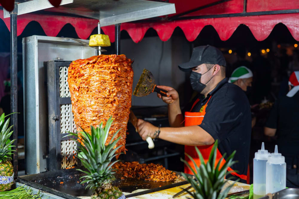 Taquero cortando carne del trompo al pastor en su puesto de la kermés