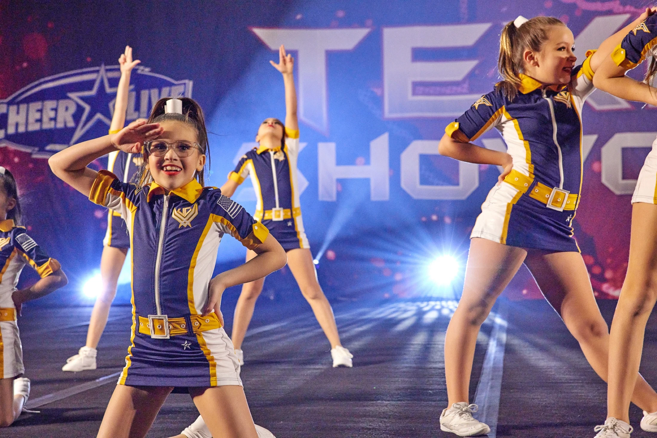 Athletes performing at Texas Showcase, with a smiling athlete in the foreground and CheerLive stage lighting behind.