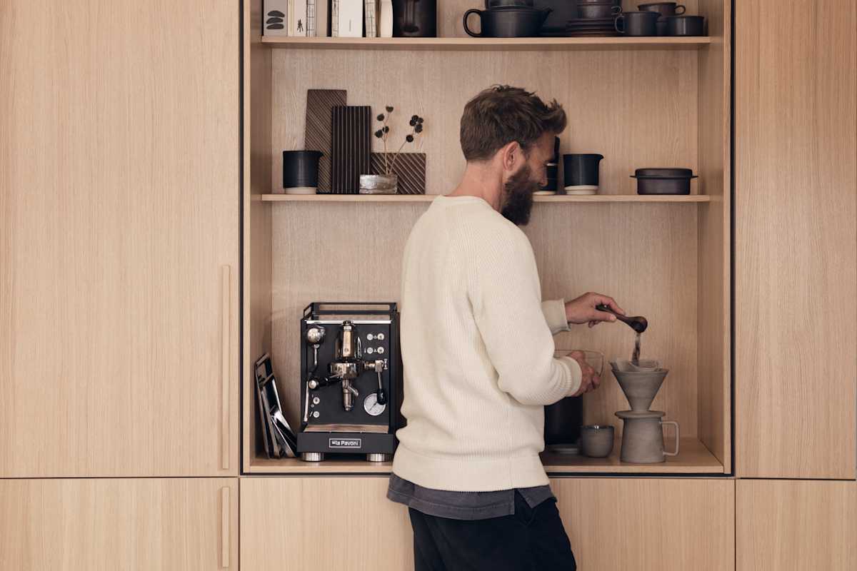 En man mäter upp kaffe i köket Ekbacken natur från Marbodal i ekfanér med vertikal träådring. Högskåp och ett öppet skåp med plats för kaffemaskin, inramad med en skuggning vilket skapar en arkitektonisk premiumkänsla.