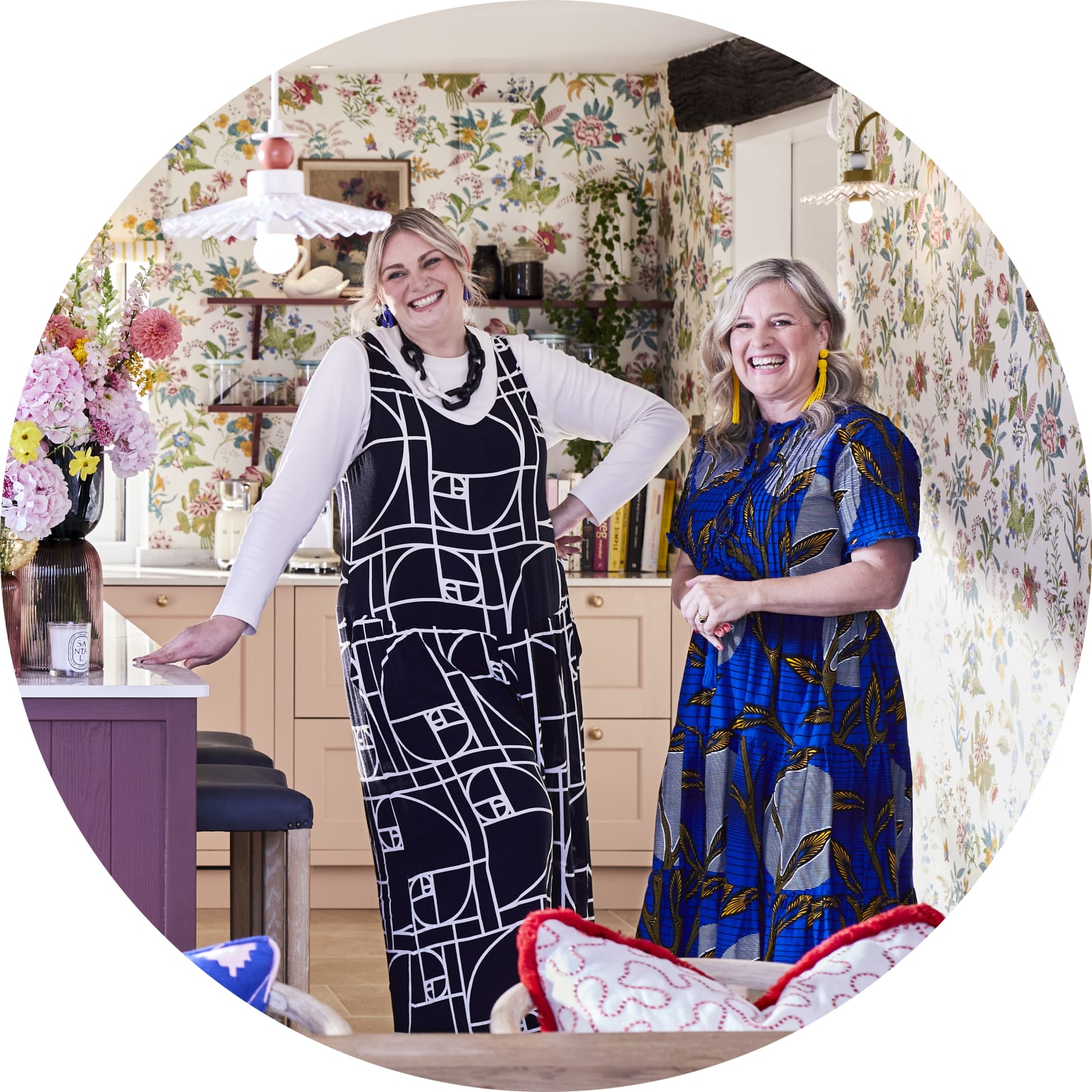 Full room view of Jen from Magnet and Sophie Robinson laughing standing next to the kitchen island in the traditional Shaker style Ludlow kitchen in colours Burlington red, Harvest and Chalk blush matched with a maximalist floral wallpaper.