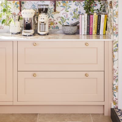 Coffee area in Sophie Robinson's maximalist kitchen, with shaker style Ludlow Chalk blush cabinetry, open shelving on a wall with a floral wallpaper and Smeg coffee machines on countertop.