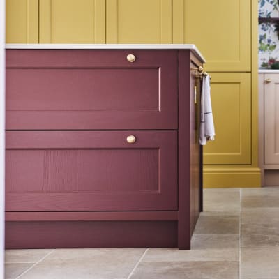 View of the corner of Ludlow Burlington Red kitchen island, Chalk Blush coloured Ludlow cabinets and Ludlow Harvest pantry in background. Brass handles, maximalist floral wallpaper and plants.