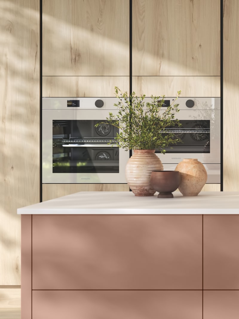 Integra Portobello Bosco as tall cabinets with integrated Samsung Ovens, white handleless Integra Soho cabinetry and a kitchen island in the shade Baked Clay, matching top cabinets and the beautiful fan above hob, all decorated with earth tone ceramics and plants.