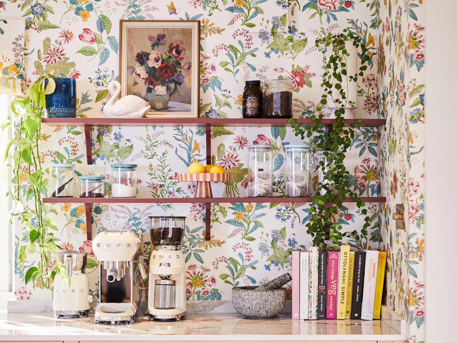 Coffee area in Sophie Robinson's maximalist kitchen, with shaker style Ludlow Chalk blush cabinetry, open shelving on a wall with a floral wallpaper and Smeg coffee machines on countertop.