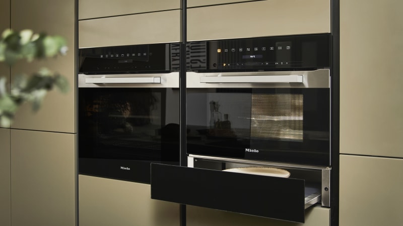 Built-in Ovens with opened warming drawer in modern slab front Balmoral Soho cabinetry from Magnet, Dekton in Ceppo worktop of kitchen island in foreground.