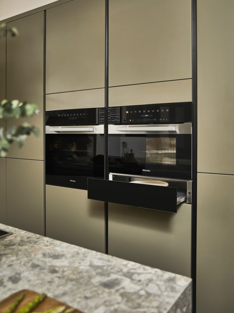 Built-in Ovens with opened warming drawer in modern slab front Balmoral Soho cabinetry from Magnet, Dekton in Ceppo worktop of kitchen island in foreground.