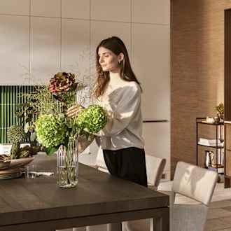 Kvinde sætter blomster i vase i mørk eg Forever Oak køkken fra INVITA i køkkenet med overskabe og højskabe i Athena Sand