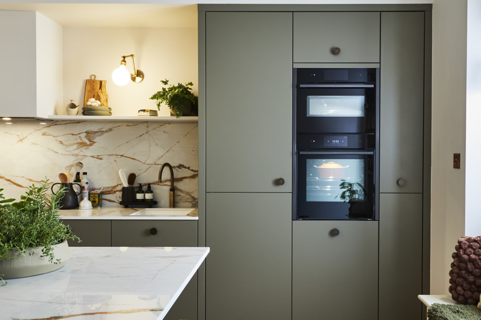 Built-in ovens in tall slab Soho Balmoral cabinets, a bench with earth tone pillows and a kitchen island with a wooden barstool and a plant