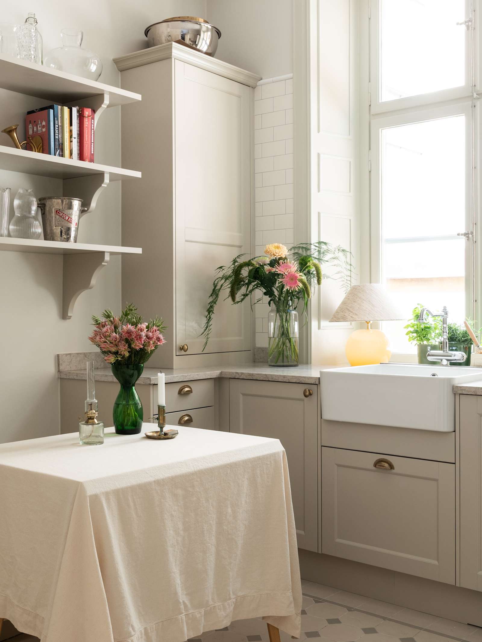 Sandra Beijers kök från Marbodal, lantliga luckan Fagerö i beige kulören Sand, med bronsfärgade handtag, diskho i vitt porslin, öppna vägghyllor i matchande beige kulör, en vit duk och mässingsljusstakar på matbordet.