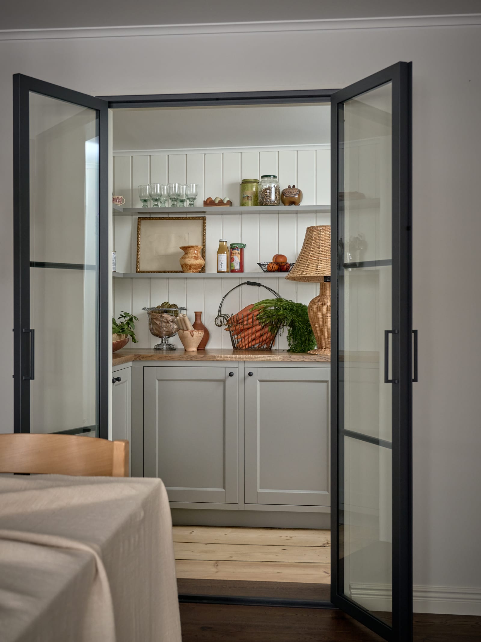 Ett walk-in skafferi med öppna glasdörrar i Malin Berghagens kök från Marbodal, lantliga Fagerö i kulören Frö med bänkskiva i trä, väggpanel bakom öppna vägghyllor och svarta knoppar.