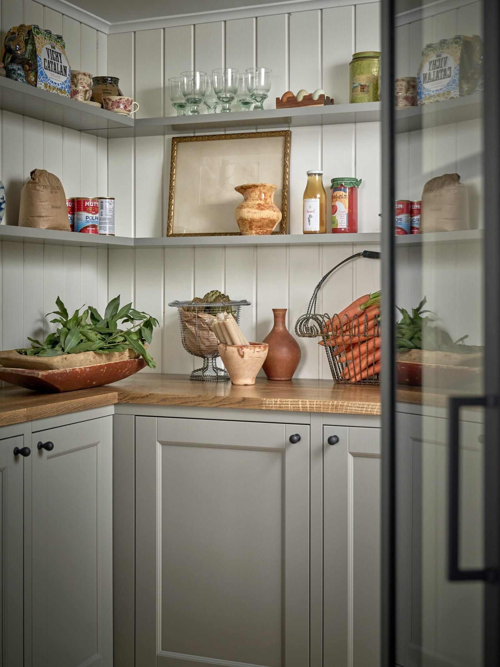 Ett walk-in skafferi med glasdörr i Malin Berghagens kök från Marbodal, lantliga Fagerö i kulören Frö med bänkskiva i trä, väggpanel bakom öppna vägghyllor och svarta knoppar.