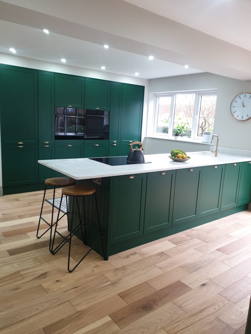 View of kitchen island and matching tall cabinets in Ashleigh's modern vibrant green Shaker Dunham, with marbled worktop and brass details like tap and cup handles.