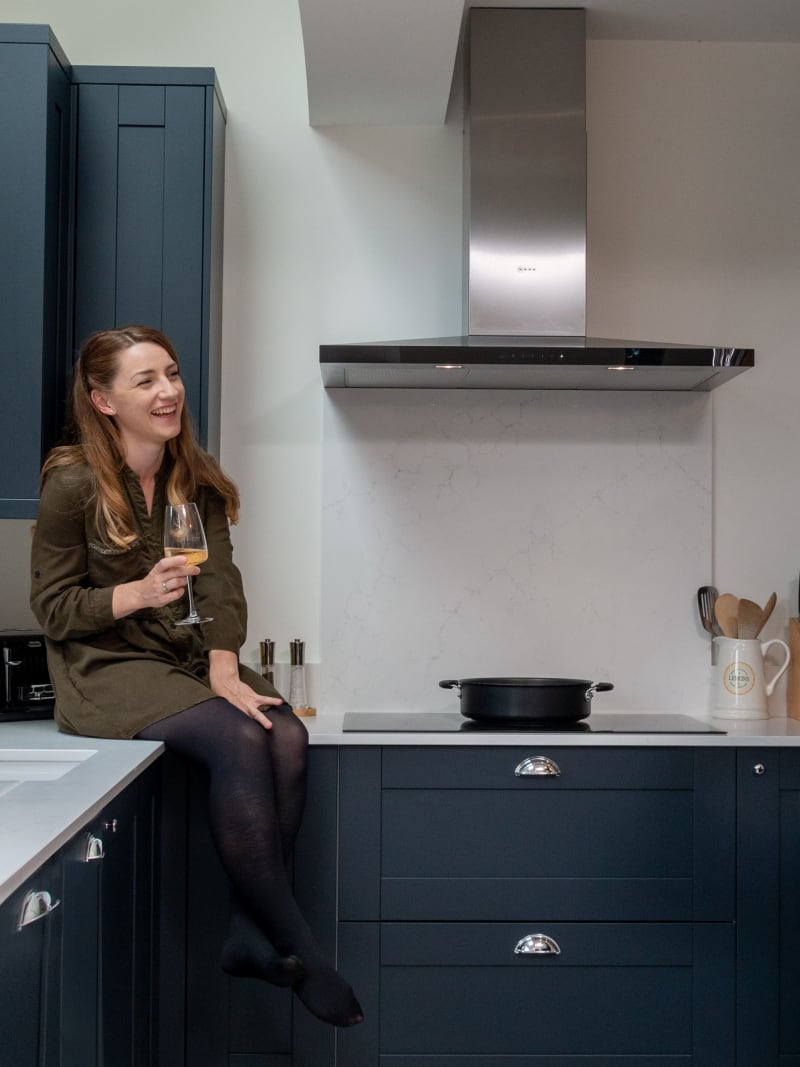 Amy sitting on the kitchen counter with a wine glass in hand while Kamal stands by the kitchen island pouring a glass of wine for himself.