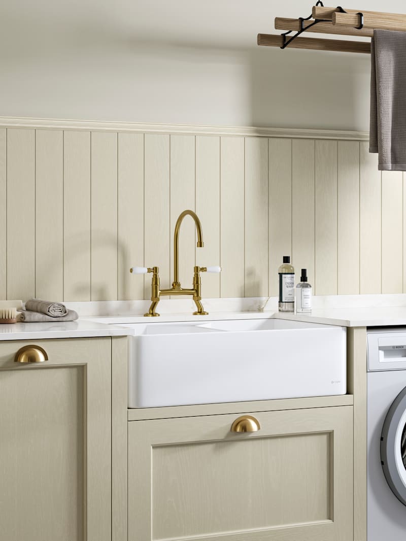 Traditional Shaker-style Ludlow Parchment and French Clay utility room with white ceramic sink, washing machine and tumble dryer with white marbled Silestone worktop and upstand, wallpanel matching the cabinetry, traditional brass tap and towels hanging on wood drying rack.