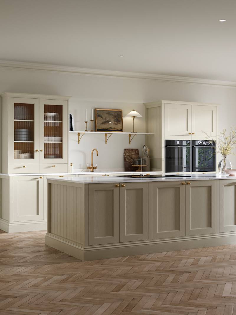 Open plan living room and Traditional Shaker-style kitchen Ludlow Parchment with tactile woodgrain, flute glass vitrine cabinets,  brass acessories like handles and tap, integrated ovens, a kitchen island and light wood herringbone flooring.