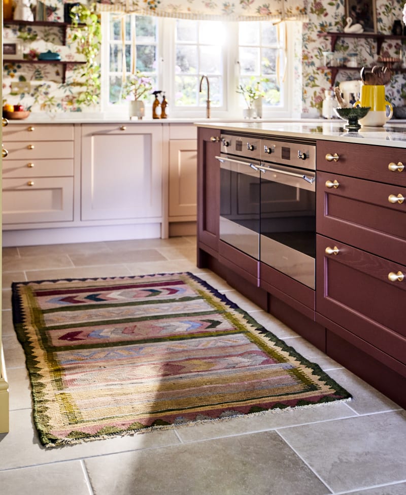 View of Ludlow kitchen island in Burlington Red, ovens in kitchen island and a woven colourful rug on the tile floor.
