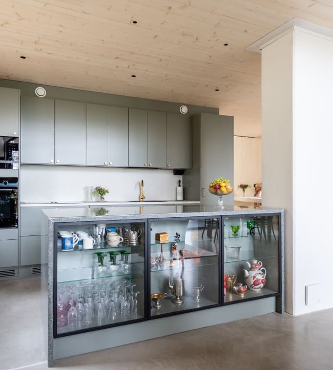 Vihreä keittiö Novartin luonnonvärisessä Havu-sävyssä, messinkinuppeilla, graniittitasoilla ja lasiovilla, joissa on alumiinikehykset keittiön altaassa.