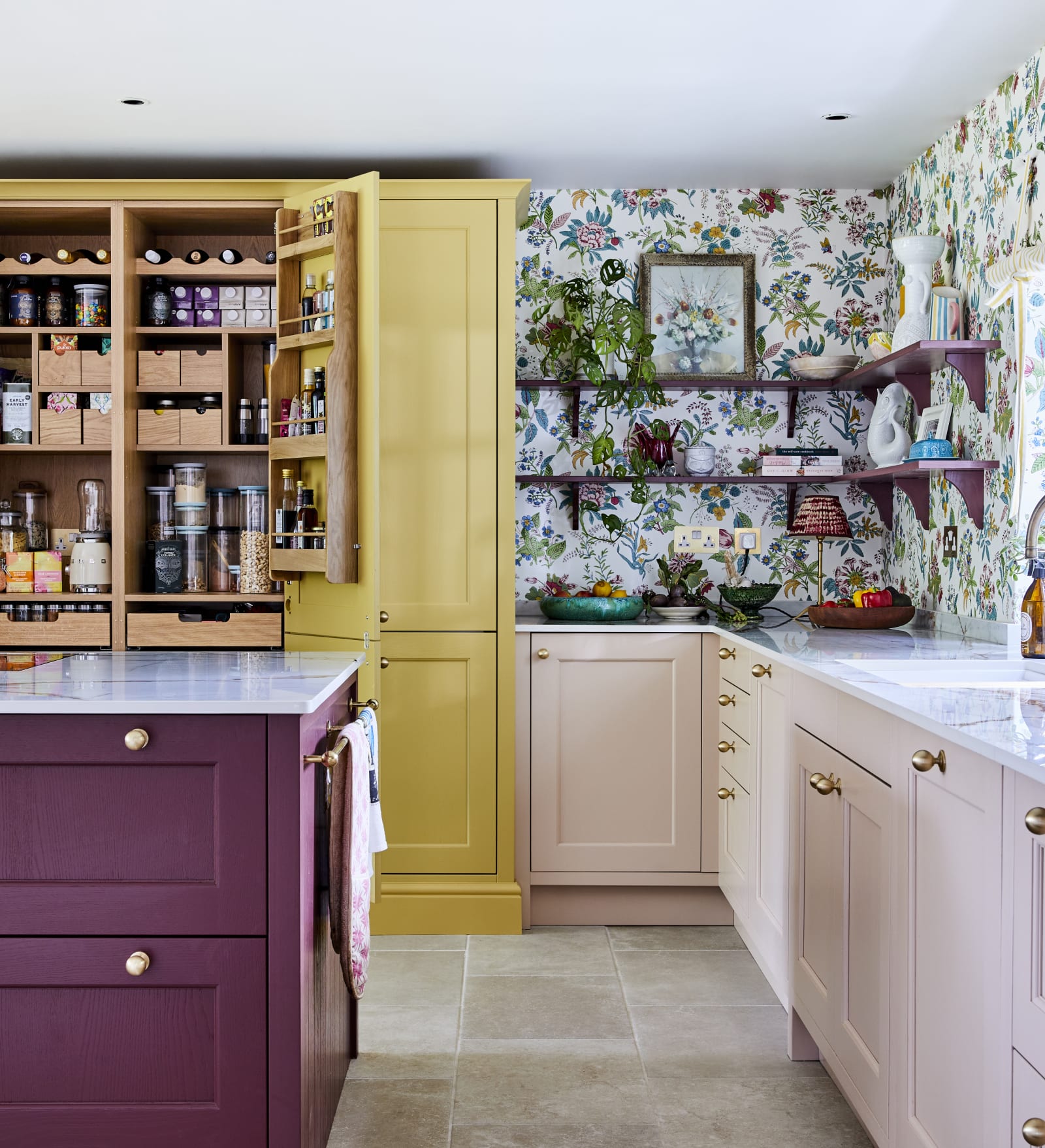 Wide image of sink, kitchen island an opened pantry larder. Ludlow cabinetry from Magnet in colours Burlington Red, Chalk blush and Harvest, wallpaper with a colourful floral pattern.