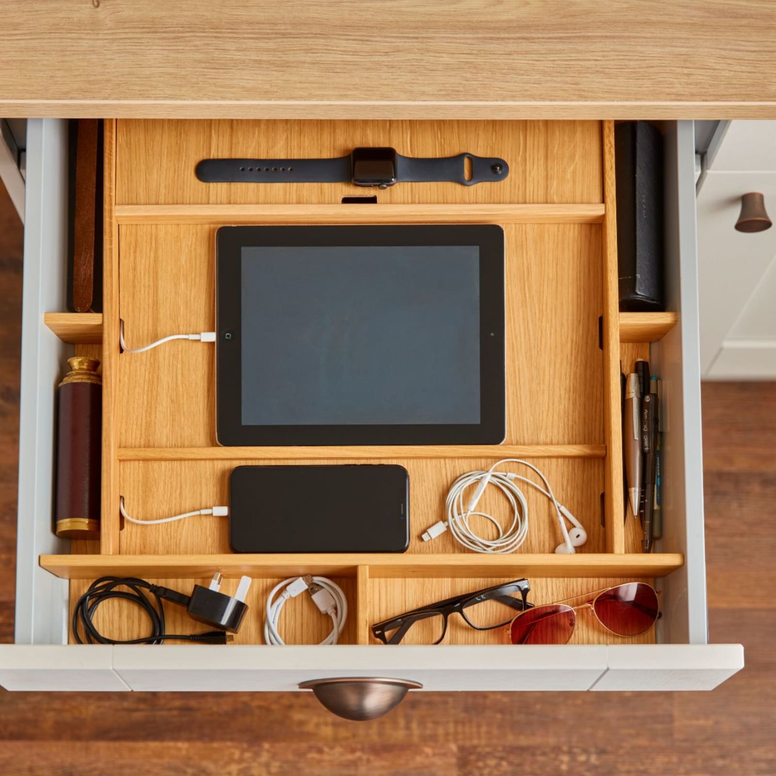 Opened drawer with oak insert holding a tablet and phone, being charged in a tray holding various electronics and glasses