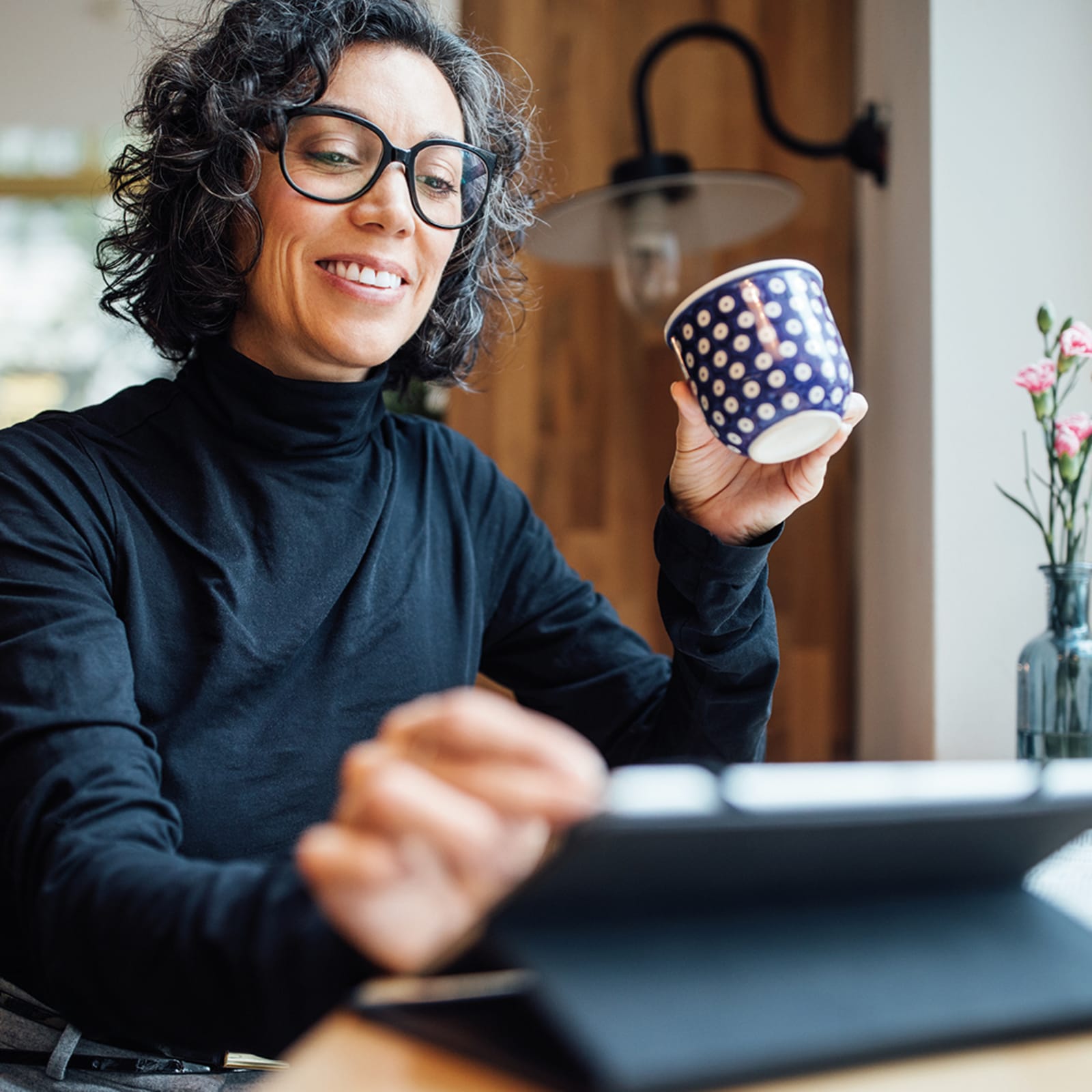 Woman wearing black roll neck smiling at tablet whilst drinking a coffee from a navy and white spotted mug