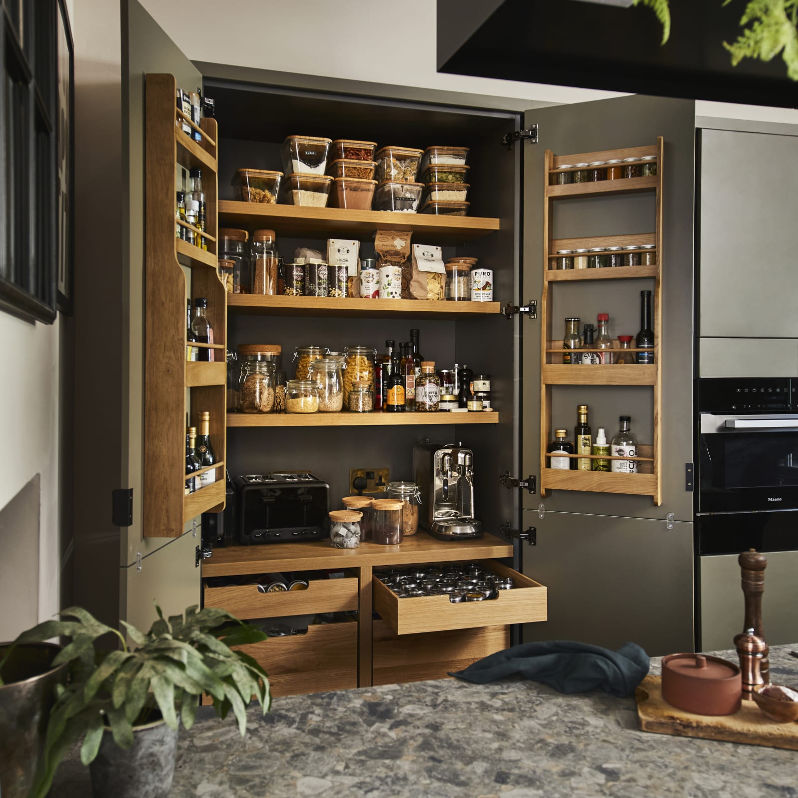 Diagonal view from kitchen island of opened pantry larder with oak interior full of glass jars containing dry goods, a toaster and spices